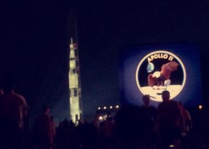 A projection of Saturn V on the Washington Monument next to a screen with a projection of the Apollo 11 mission seal